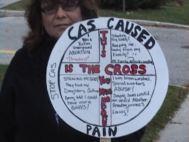 Aboriginal woman with sign CAS Caused Pain, This is the cross you made me bare